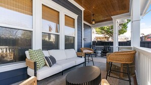 Ein überdachter Balkon mit einem weißen Sofa, Korbstühlen, einem runden Tisch und einer Holzbeschichtung an der Decke.