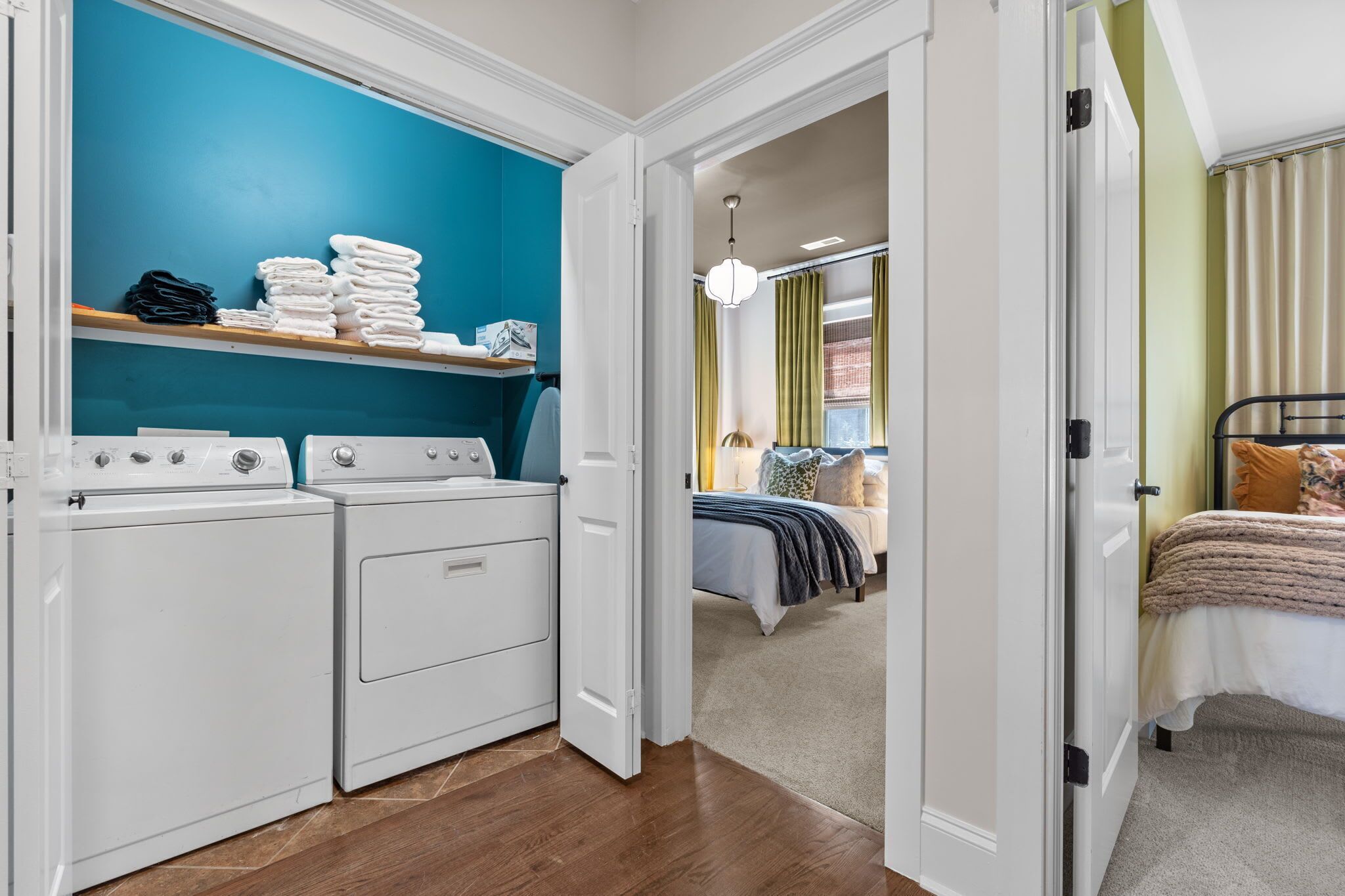 A laundry area with stacked towels and a washing machine, a bedroom with a bed and a nightstand, and a hallway leading to another room.