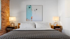 A neatly made bed with a grey knitted blanket, two bedside tables with lamps, a brick wall, and a large abstract painting on the wall. - OneTreeHill Loft Downtown, Riverwalk, Live Oak (Wilmington)