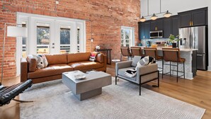 A spacious living area with a brick wall, a brown leather sofa, a grey armchair, and a kitchen with dark cabinets and stainless steel appliances. - OneTreeHill Loft Downtown, Riverwalk, Live Oak (Wilmington)