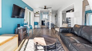 Un salon moderne avec un canapé en cuir noir, une table basse en bois, un téléviseur fixé au mur et un ventilateur de plafond.