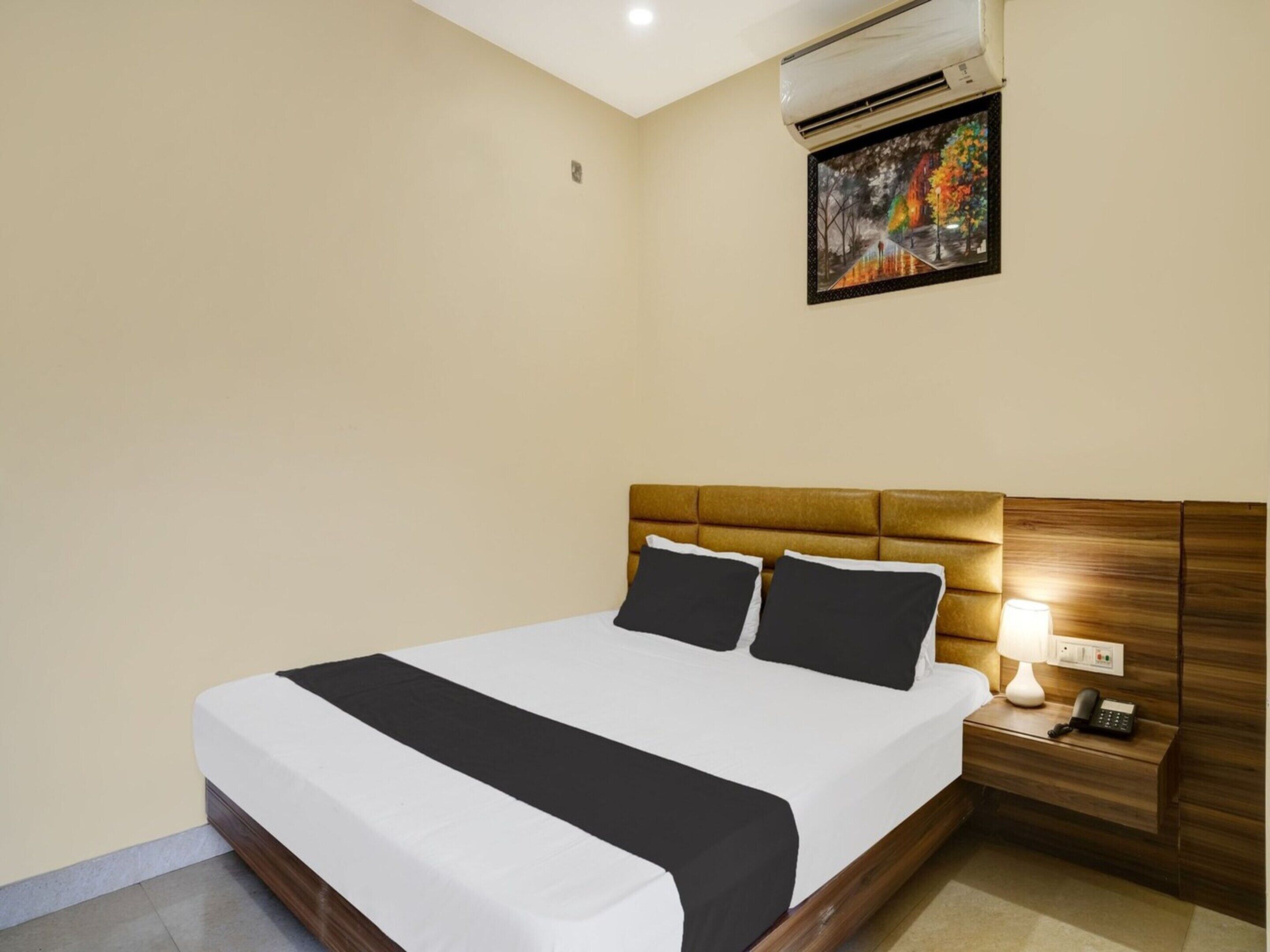 A hotel room with a bed, a nightstand with a lamp and phone, and an air conditioning unit.