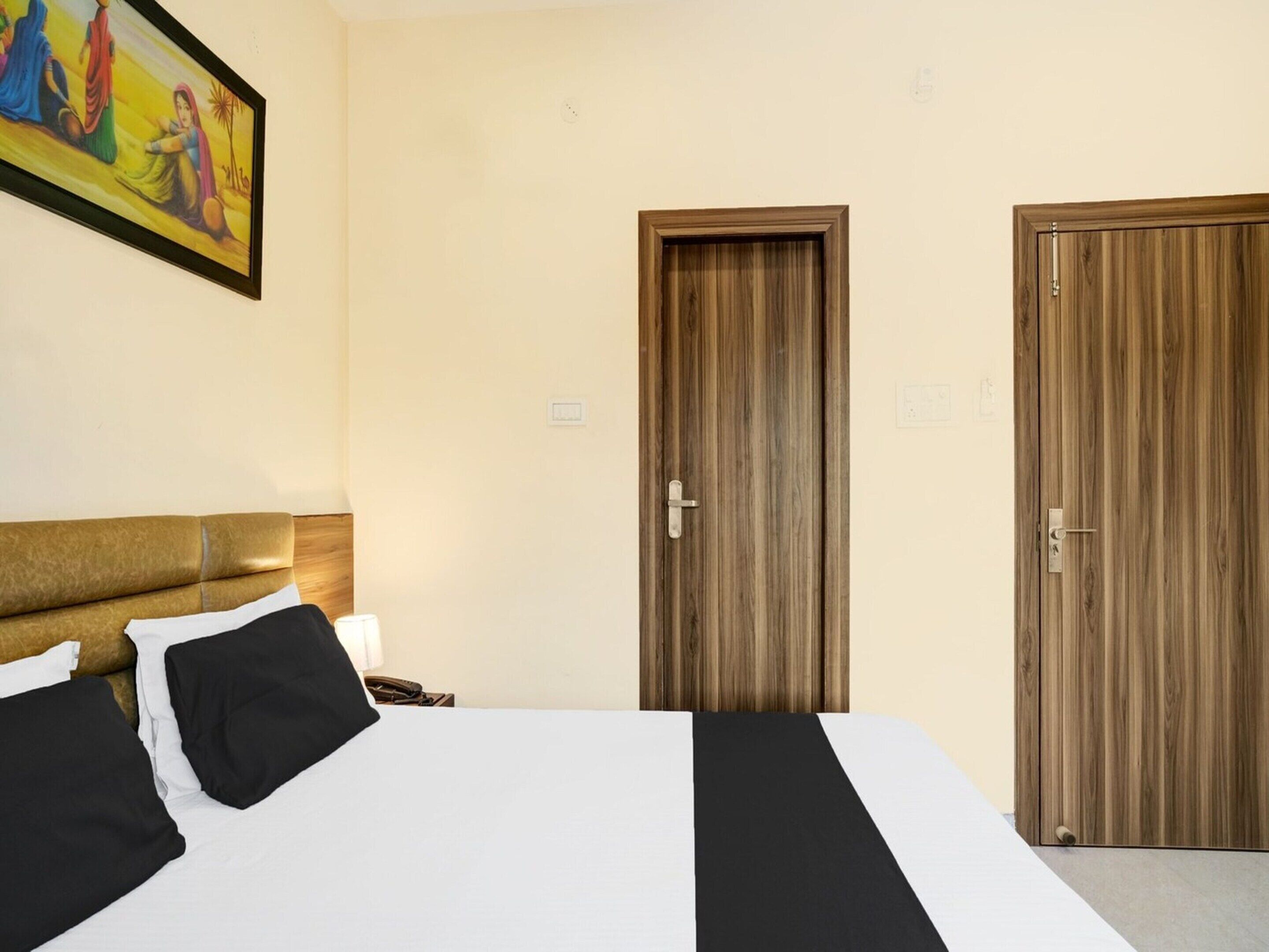 A hotel room with a bed, two doors, a painting on the wall, and a nightstand with a lamp.