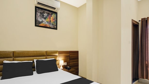 A hotel room with a large bed, a headboard, an air conditioning unit, a painting on the wall, a bedside lamp, and a door with curtains. - Deluxe Lalganesh Formally Royal Avenue (Guwahati)
