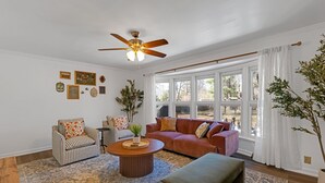 Un salon avec un ventilateur de plafond, un canapé, un fauteuil, une table basse et une grande fenêtre avec des rideaux.