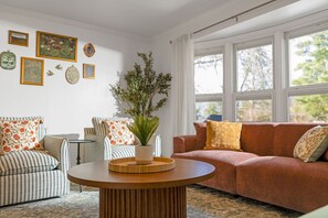 A living room with a striped sofa, a round wooden coffee table, a red sofa, and a large window with curtains.