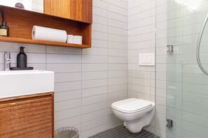 A modern bathroom with a toilet, a wooden shelf with toiletries, and a glass shower enclosure.