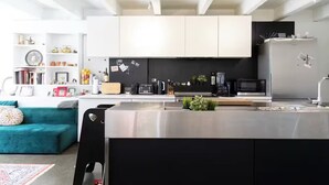 Une cuisine moderne avec des meubles blancs, un plan de travail noir et des appareils en acier inoxydable. Le salon est agrémenté d’un canapé turquoise et d’un fauteuil noir.