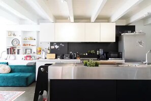 A modern kitchen with white cabinets, a black countertop, and stainless steel appliances. There is a teal sofa and a black chair in the living area. - Walk to Everything! Bright & Stylish Fully Furnished Stay (Montréal)