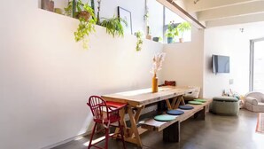 Un coin repas avec une table en bois, des bancs et des chaises rouges. Des plantes en pot sont disposées sur le mur, et un téléviseur y est fixé.