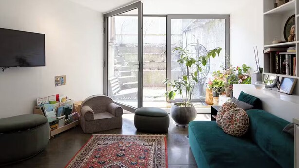 Un salon comprenant un canapé, un fauteuil, un pouf et un téléviseur. On y trouve également des plantes en pot et une bibliothèque.