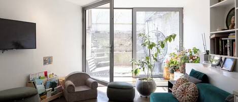 A living room with a sofa, armchair, ottoman, and a television. There are potted plants and a bookshelf.