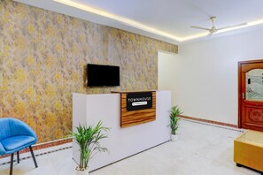 A reception desk with a sign, a flat-screen TV, a ceiling fan, a blue chair, and two potted plants. - Single Room Madiwala Lake Park (Bengaluru)
