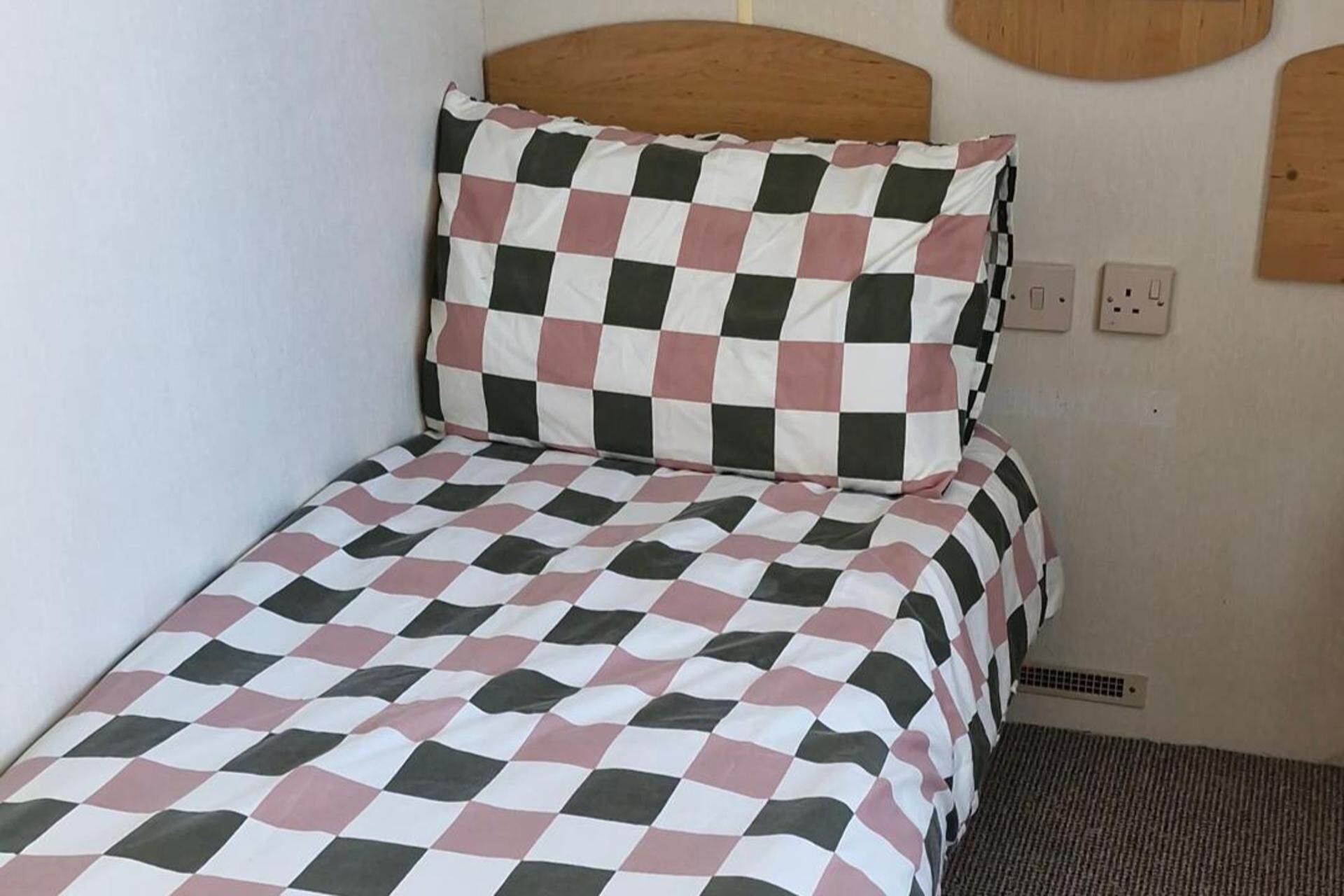 A single bed with a checkered bedspread, a wooden headboard, and a wall socket.