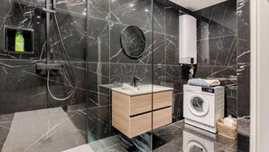 Ett modernt badrum med en duschavdelning med glasväggar, ett badrumsskåp i trä, en inbyggd tvättmaskin och en vägg beklädd med marmorplattor.