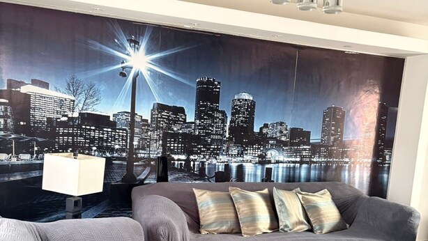 Un salon avec une fresque murale représentant un paysage urbain, un canapé gris, une table basse en verre et une lampe.