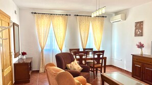 Ein Wohnzimmer mit einer Couch, Sesseln, einem Tisch, einem Schrank und einem Fenster mit Vorhängen.