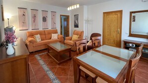 Un saló amb un sofà, dues butaques, una taula de centre, una taula de menjador i una televisió. Hi ha quadres amb marc a la paret i una porta de fusta.