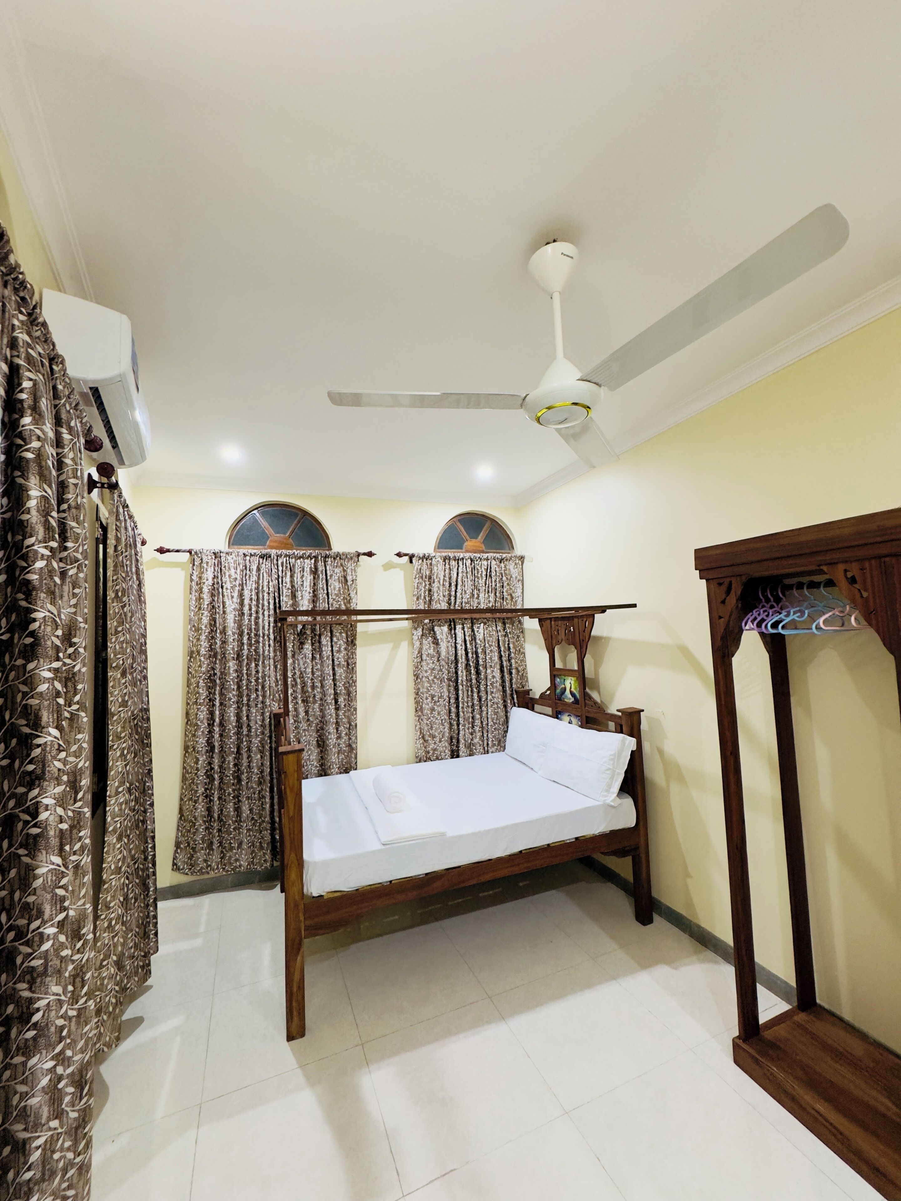 A bedroom with a bed, ceiling fan, air conditioning unit, curtains, and a wooden wardrobe.