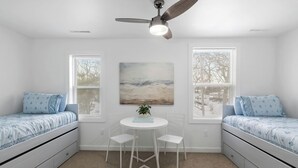 Habitación con dos camas, ventilador de techo, un cuadro, una mesita con sillas y ventanas con persianas.