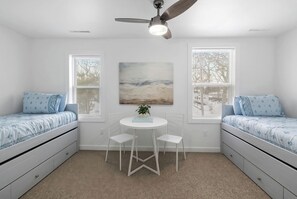 Une chambre avec deux lits, un ventilateur de plafond, un tableau, une petite table avec des chaises et des fenêtres équipées de stores.