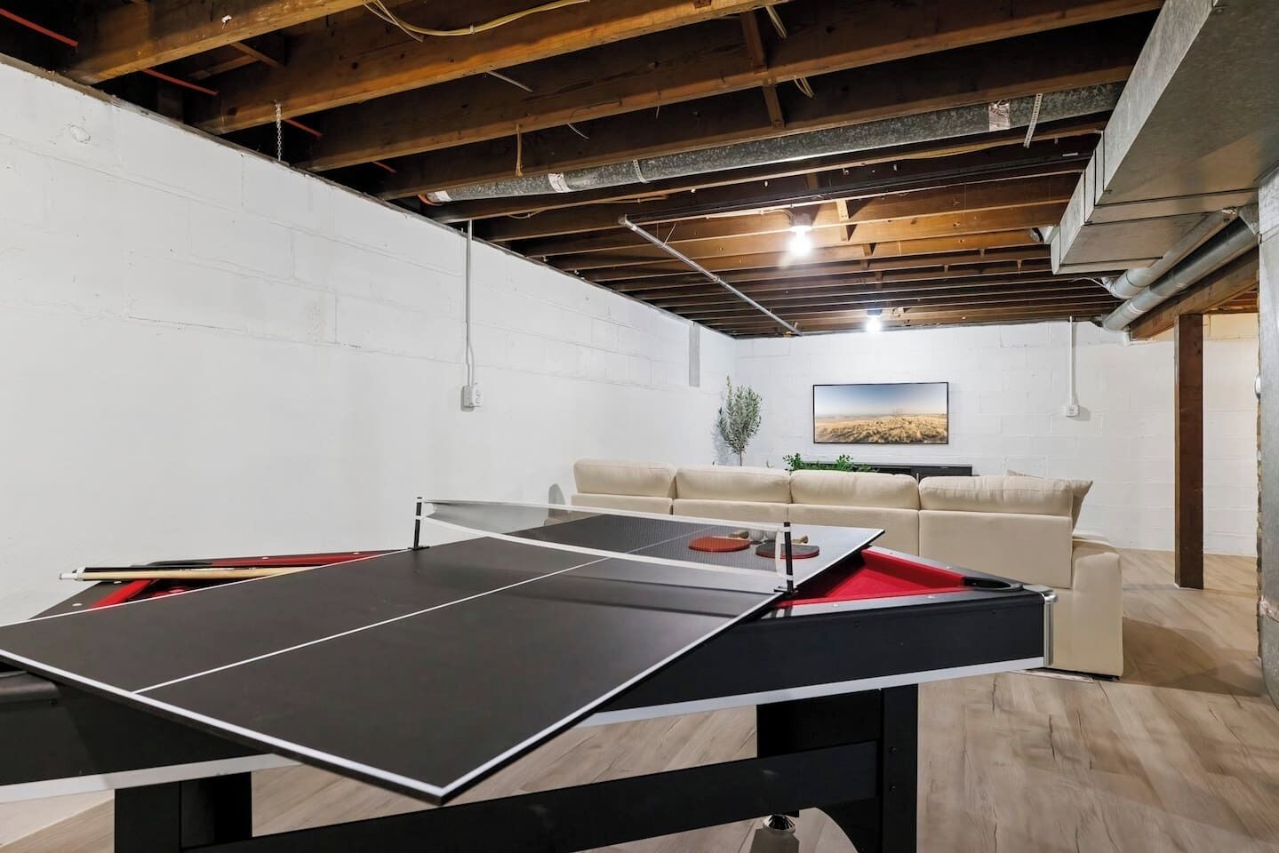 A ping pong table in a room with a white brick wall, a beige sofa, and a painting on the wall.