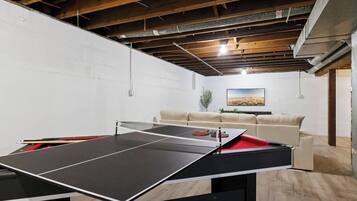 A ping pong table in a room with a white brick wall, a beige sofa, and a painting on the wall.