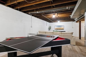 Una mesa de ping pong en una habitación con pared de ladrillo blanco, un sofá beige y un cuadro en la pared.