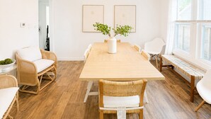 Comedor con mesa de madera, sillas de mimbre, un florero blanco con plantas y una lámpara colgante.