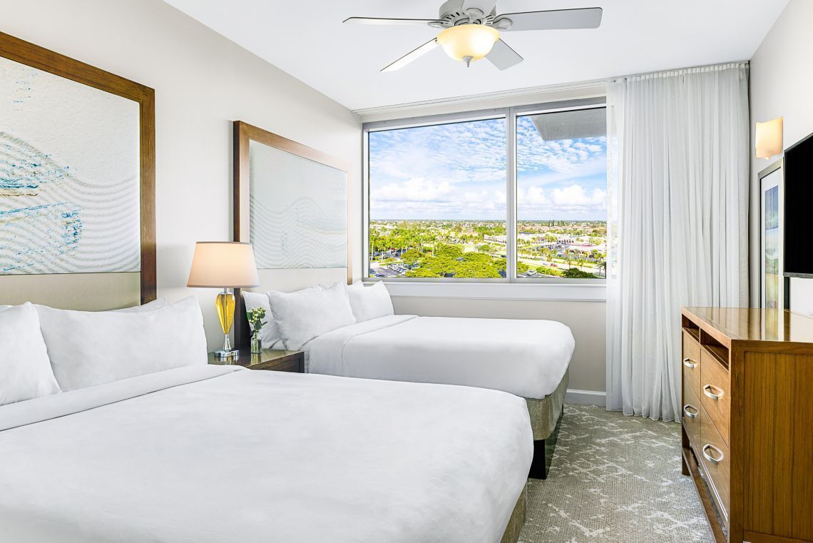 A hotel room with two beds, a ceiling fan, a large window with a city view, and a wooden dresser.