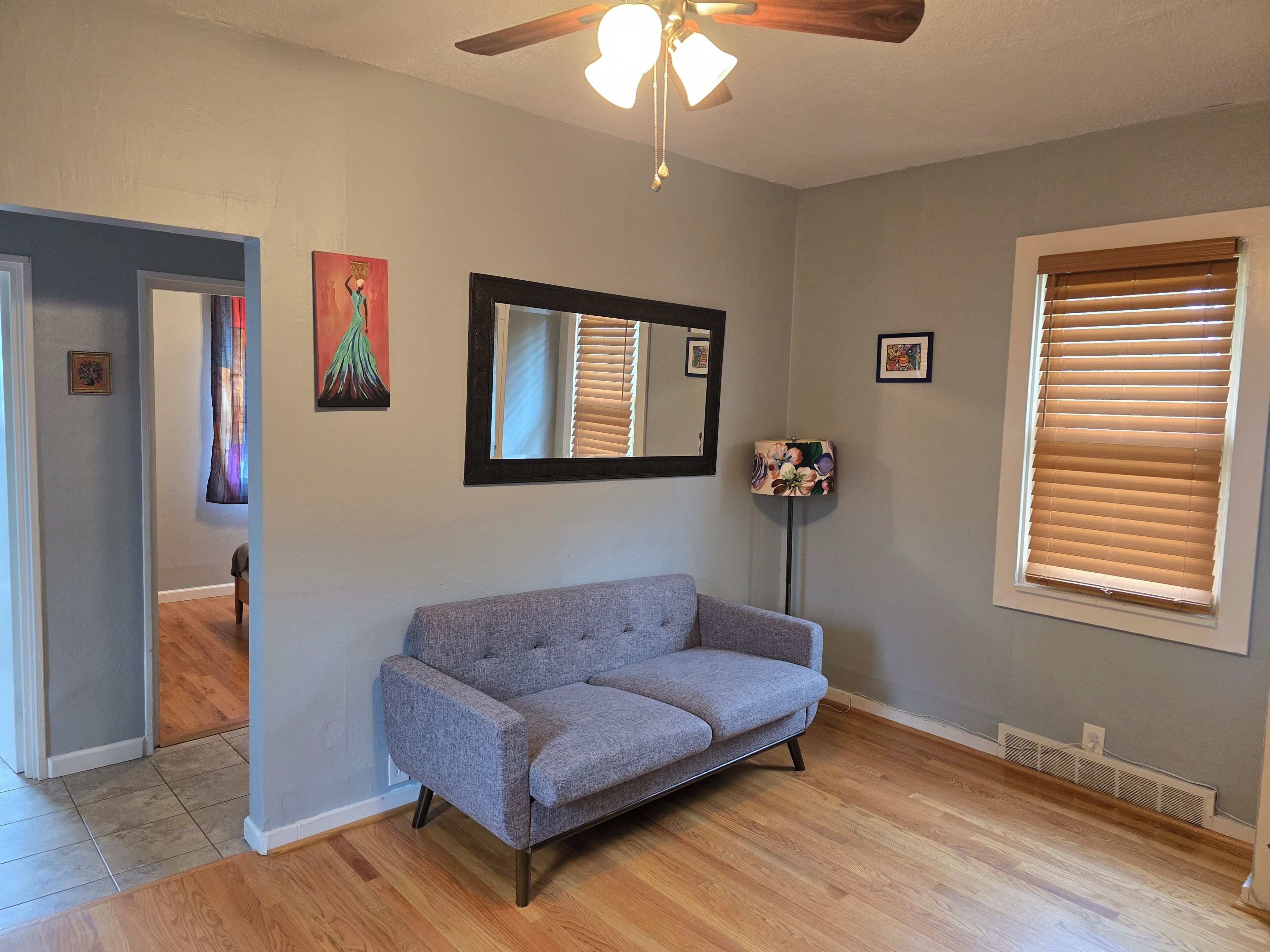 A room with a gray sofa, a ceiling fan, a mirror, and a window with blinds.