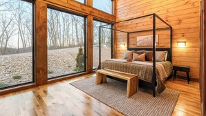 A wooden cabin bedroom with a four-poster bed, a bench, and a large window offering a view of a gravel path and trees. - Luxury Smoky Mountain Cabin w/ Sauna (Sevierville)