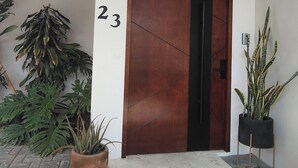 Une entrée d’immeuble avec une porte numérotée, des plantes en pot et un chemin en béton.