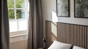 Un dormitorio con cama, ventana con cortinas, radiador, mesita de noche con lámpara, y dos cuadros en la pared.