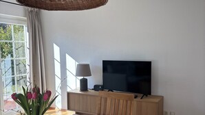 Un área de comedor con una mesa de madera, sillas, un florero con flores, un televisor sobre un mueble de madera y una lámpara colgante de mimbre.