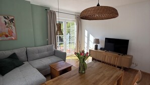 Una sala de estar con sofá, mesa de comedor de madera, sillas, televisor y una ventana con cortinas.
