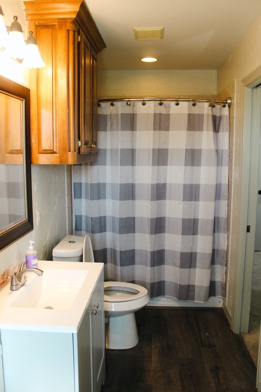 A bathroom with a checkered shower curtain, a toilet, a sink with a mirror, and wooden cabinets.