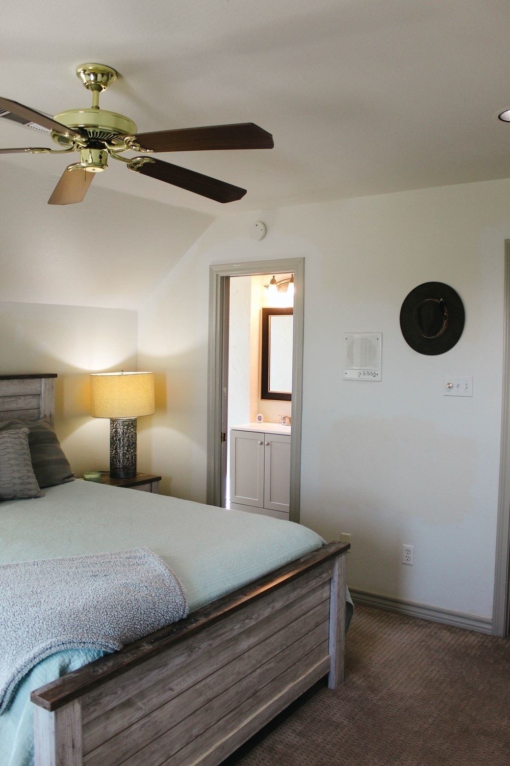 A bedroom with a bed, a ceiling fan, a nightstand, a lamp, a door, and a wall-mounted hat.