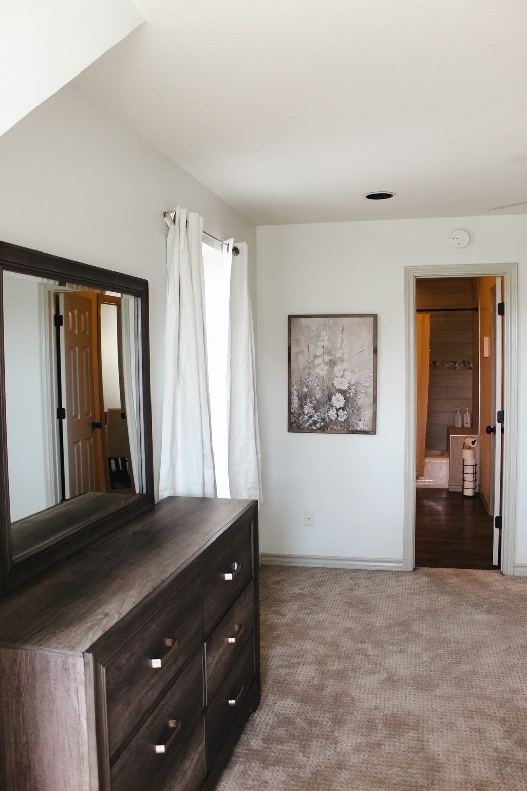 Une chambre avec une commode en bois foncé, un grand miroir, un rideau blanc, une œuvre d’art florale encadrée et une porte donnant sur une salle de bain.