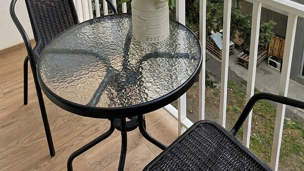 A balcony with a round glass table, two wicker chairs, a white railing, and a view of trees and buildings. - Brand New - 70 m² - Family Friendly - Fast WiFi - 5 min to Old Town (Tbilisi)