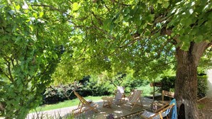 A covered outdoor seating area with wooden chairs and a table, surrounded by trees and greenery. - Drôme provençale house with swimming pool and river (La Bégude-de-Mazenc)
