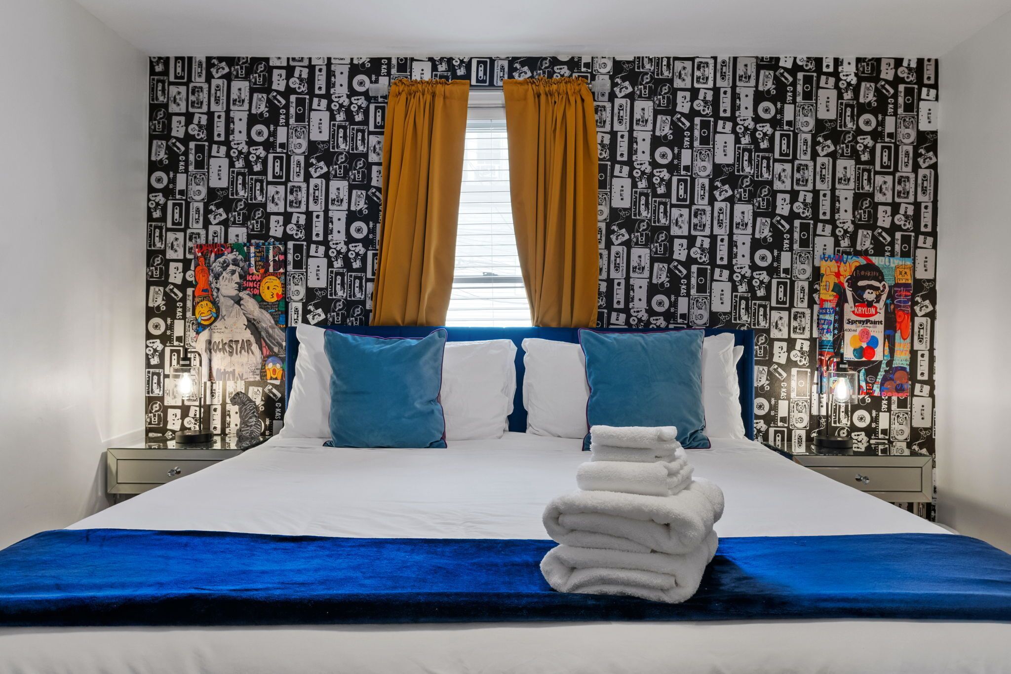 A hotel room with a bed, pillows, a stack of towels, and a wall with a cassette tape pattern.