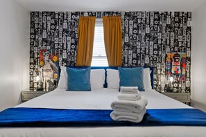 A hotel room with a bed, pillows, a stack of towels, and a wall with a cassette tape pattern. - Modern Lawrenceville 4BR w/ Deck + 2 Car Garage w/ EV Charger - Sleeps 11 (Pittsburgh)