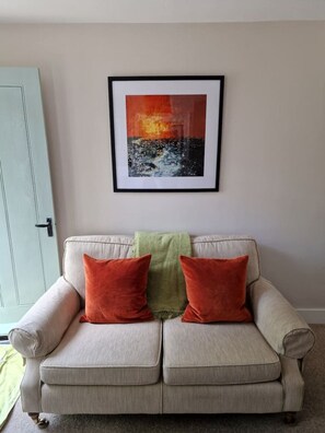 Ein beiges Sofa mit zwei roten Kissen und einer grünen Decke, darüber ein gerahmtes Bild und links eine hellblaue Tür.