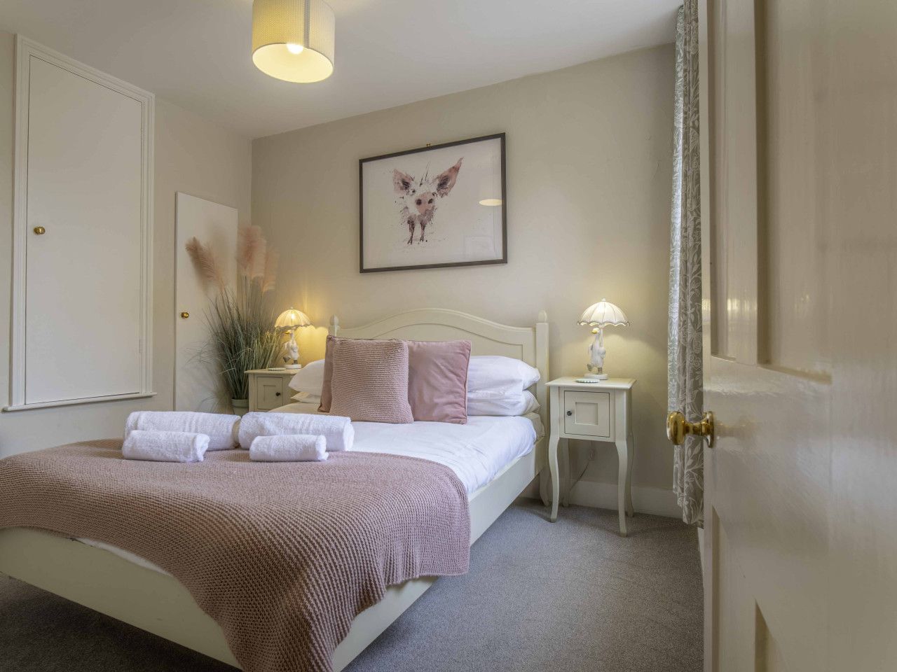 A neatly made bed with pillows and a blanket, a bedside table with a lamp, a framed picture on the wall, and a door with a curtain.