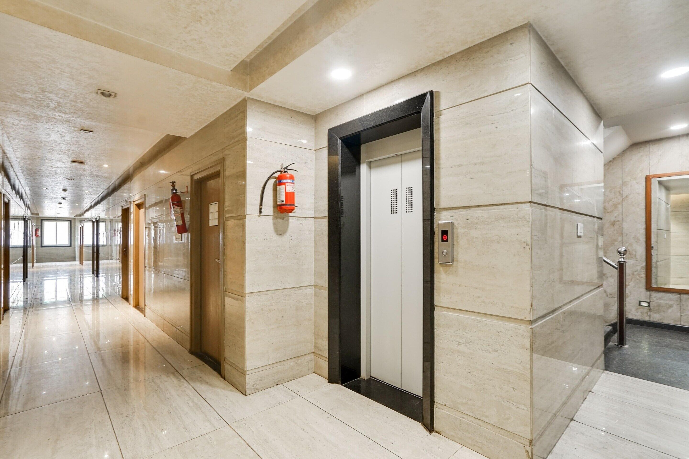 A modern elevator with a digital display, a fire extinguisher mounted on the wall, and a polished marble floor in a hallway with doors on the left.