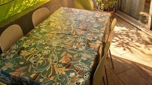 A dining area with a table covered by a patterned tablecloth, surrounded by chairs, and a yellow curtain partially drawn. - Beautiful Apartment in Residence with Swimming Pool, Saint-Cyprien. (Saint-Cyprien)