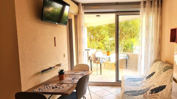 A small room with a dining table, chairs, a sofa, a TV mounted on the wall, and a sliding glass door leading to an outdoor area with a table and chairs. - Beautiful Apartment in Residence with Swimming Pool, Saint-Cyprien. (Saint-Cyprien)
