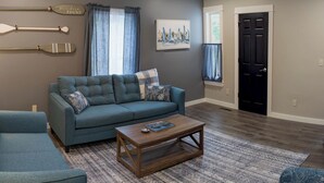 A living room with a blue sofa, a wooden coffee table, and a rug. There are two windows with curtains, a painting on the wall, and a door. - Modern NewlyFurnished Acadia Apt (Ellsworth)