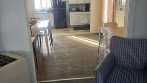 Een woonkamer met een gestreept fauteuil, een eettafel met stoelen, een keuken met kasten, en een plafondventilator.